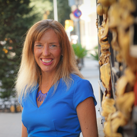 Kat O'Brien smiles as she stands next to a brick wall in a blue dress for her headshot