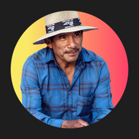 A hispanic man smiles wearing a blue plaid shirt and hat that says "Made in Mexico."