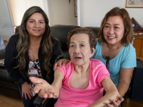 An elderly mother reaching out to the camera, next to her caretaker daughter and their caregiver navigator