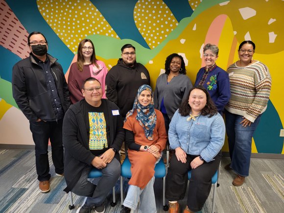 A group photo of the Wellspring grantmaking committee, 9 people smiling at the camera in front of a colorful mural