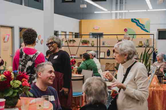 Photo of various groups of people sitting and standing in conversation around orange tables with bright flower arrangements.