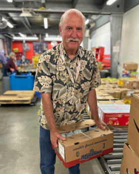 David Bayer smiling and holding a box of food while volunteering in Food Share's warehouse